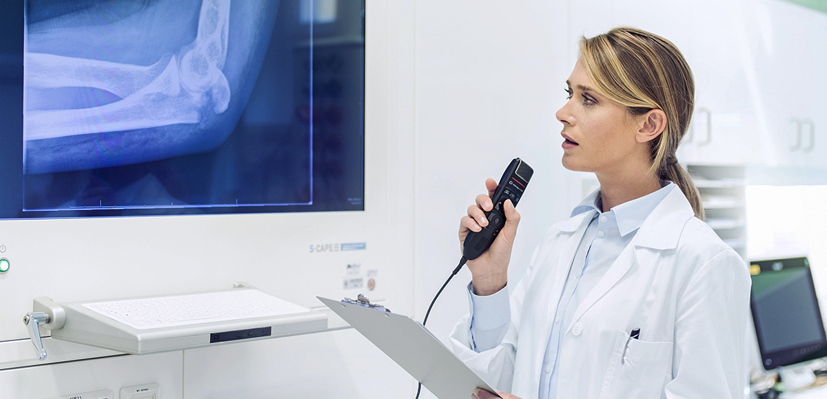 A woman in a white coat, likely a doctor or researcher, speaking into a dictation device. She is examining an x-ray, surrounded by medical equipment in an indoor setting, possibly within a research institute or laboratory. The scene highlights her role in the healthcare field and her engagement with medical technology.