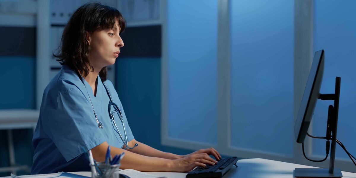 Eine medizinische Fachkraft in einem blauen Bluse sitzt konzentriert vor dem Computer und tippt auf der Tastatur. Die Frau wirkt erschöpft.