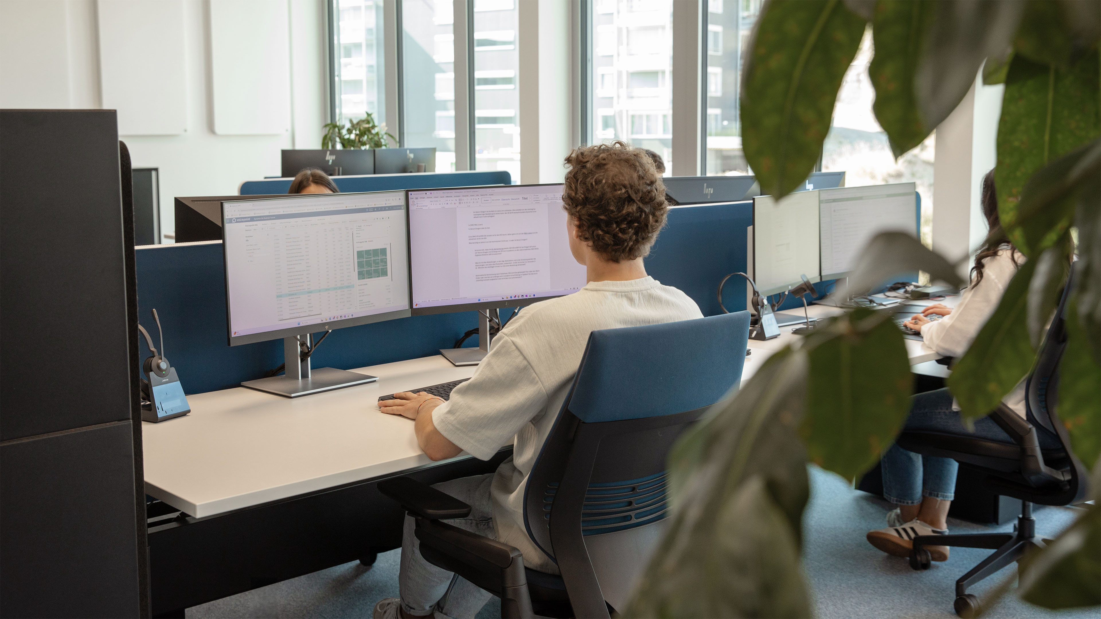 Several employees sit at their desks in an open-plan office, concentrating on their work on two screens each.