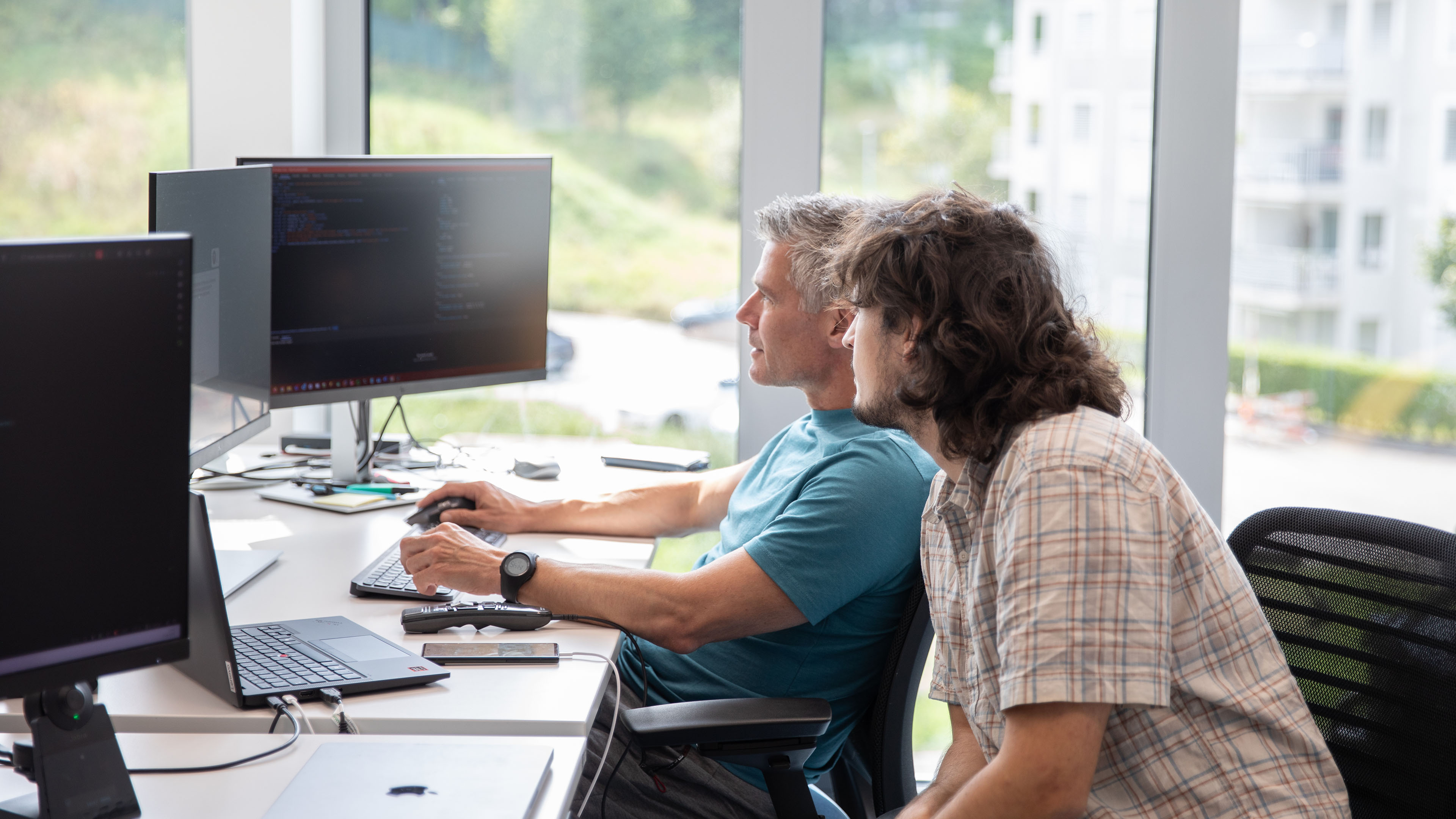 Two employees sit at their workstations in front of two screens and work together on a software solution.