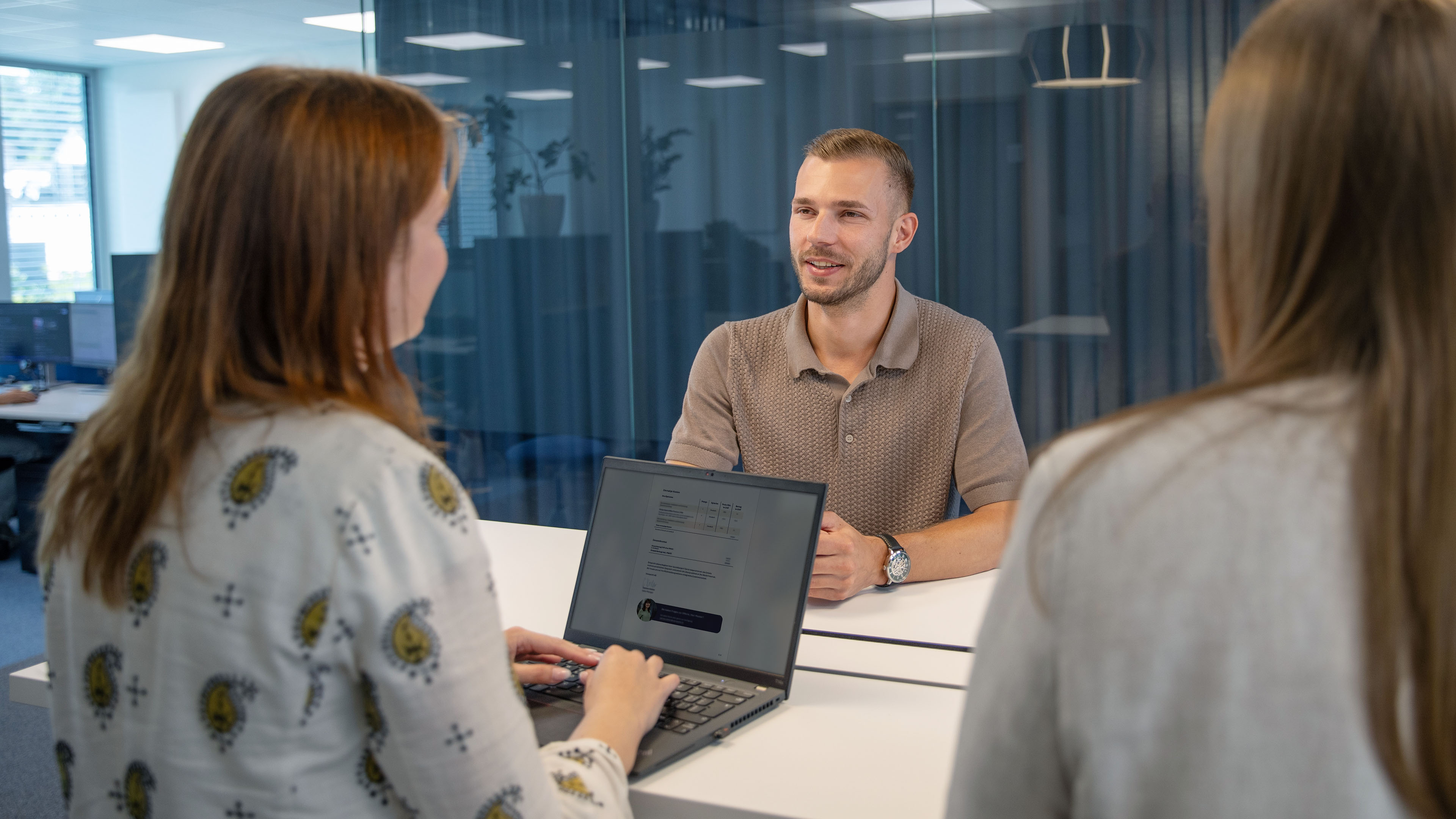 Three employees are sitting at a table having a conversation, while one person is working on a laptop.