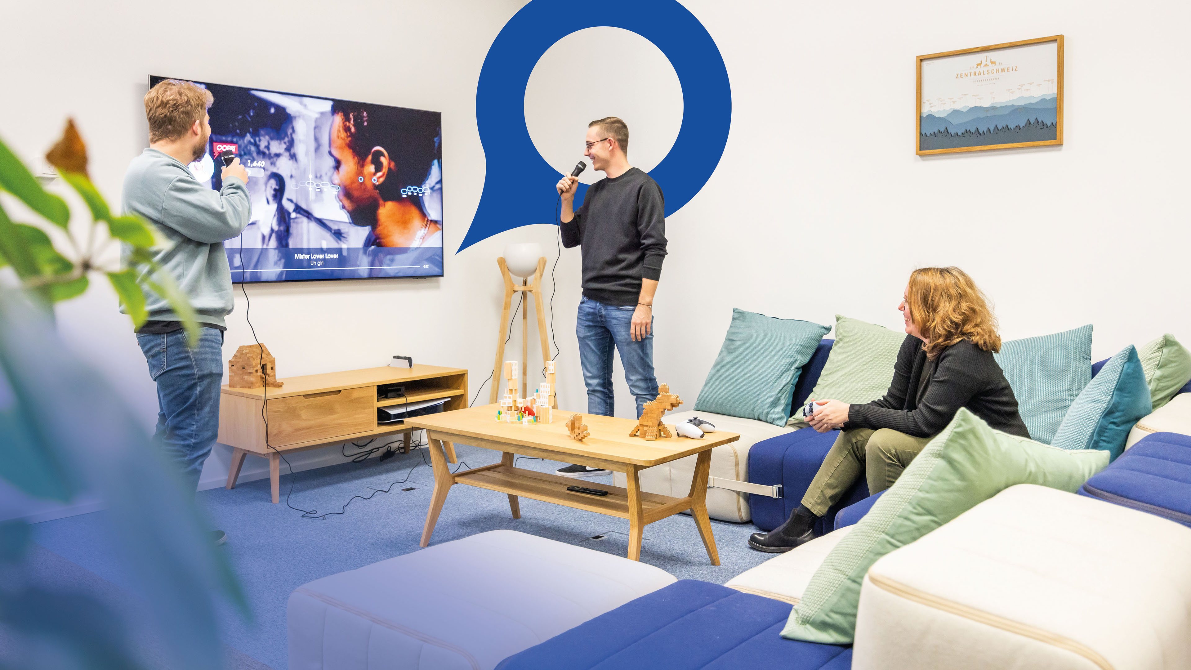 Zwei Mitarbeitende stehen im Aufenthaltsraum des Voicepoint-Büros in Wetzikon und singen Karaoke. Im Hintergrund ist die blaue Voicepoint-Sprechblase sichtbar.