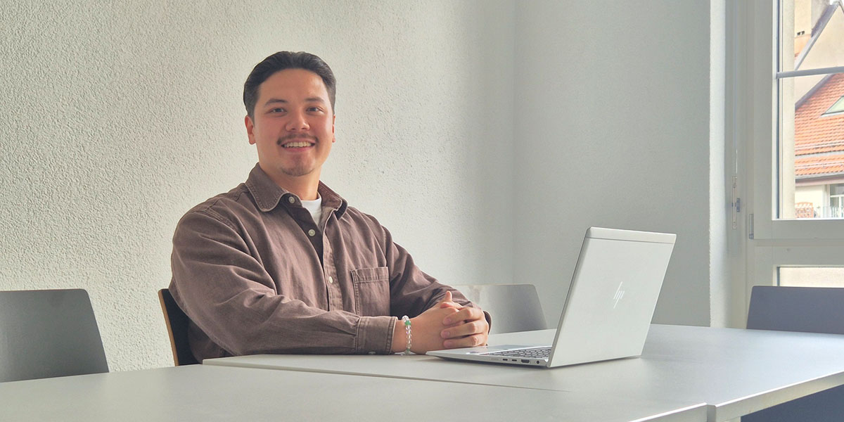 Un homme est assis à une table blanche, un ordinateur portable ouvert devant lui, et sourit à la caméra. Il porte une chemise marron sur un t-shirt blanc.