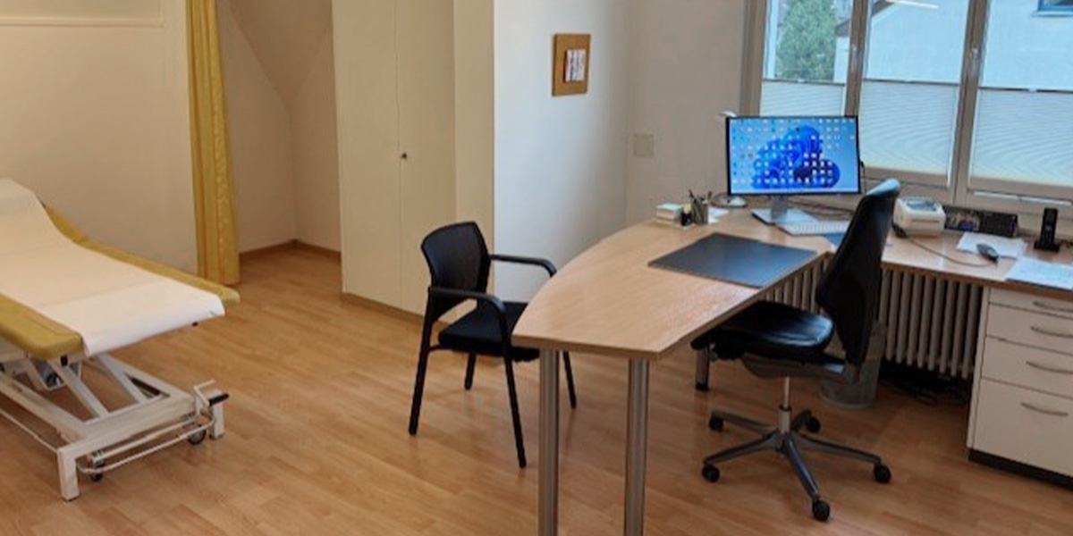 Aufnahme eines Behandlungszimmer. Rechts befindet sich ein Schreibtisch mit zwei schwarzen gegenüberstehenden Stühlen und links ist ein gelbes Bett mit einem weissen Tuch zu sehen.