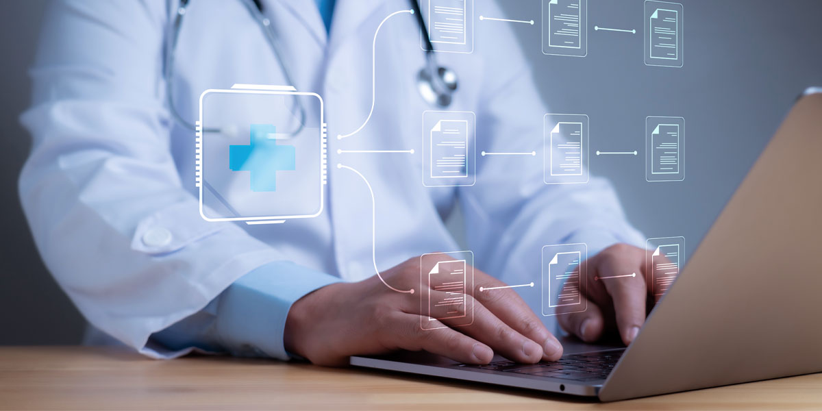 A doctor in a white coat sits at a desk and enters information into an electronic health record on a computer.
