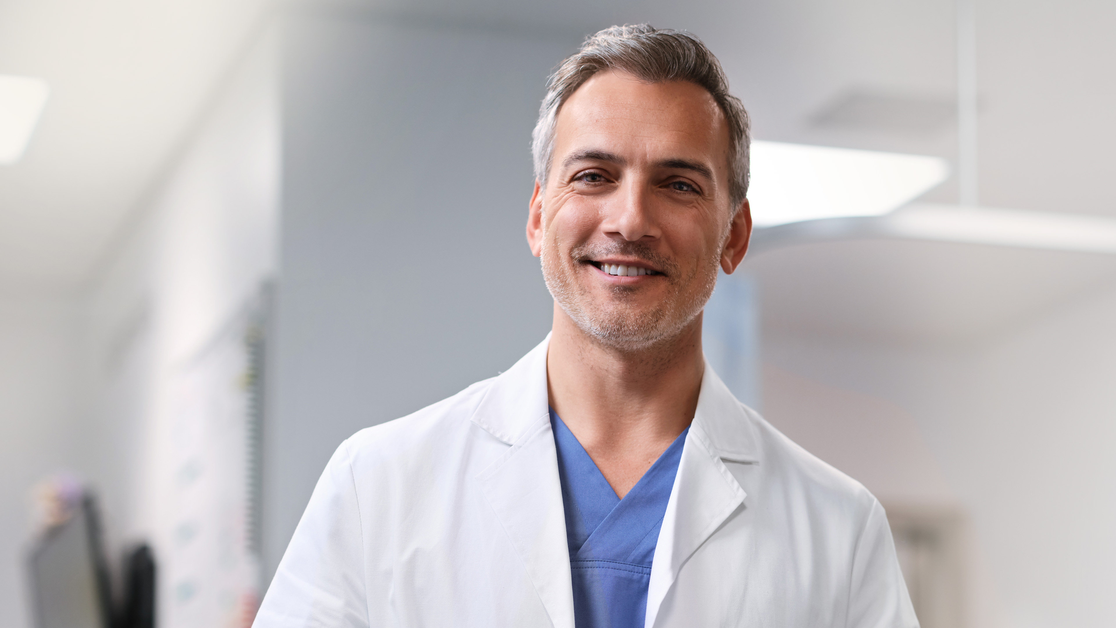 Ein Arzt im weissen Kittel steht in einem Klinikgang und lächelt in die Kamera