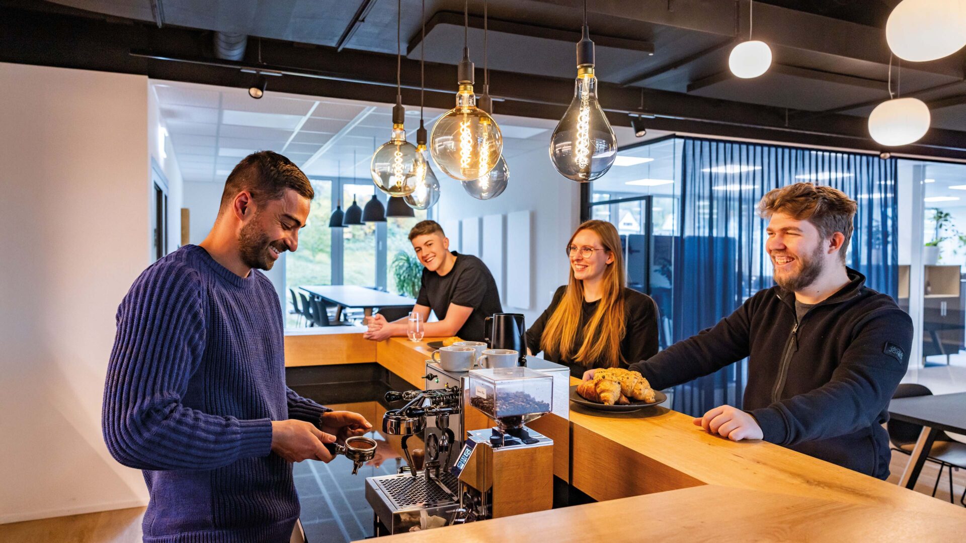 Drei Mitarbeitende sind in der Cafeteria des Voicepoint-Büros in Wetzikon. Zwei sitzen an der Bar, eine Person steht dahinter und macht Kaffee.