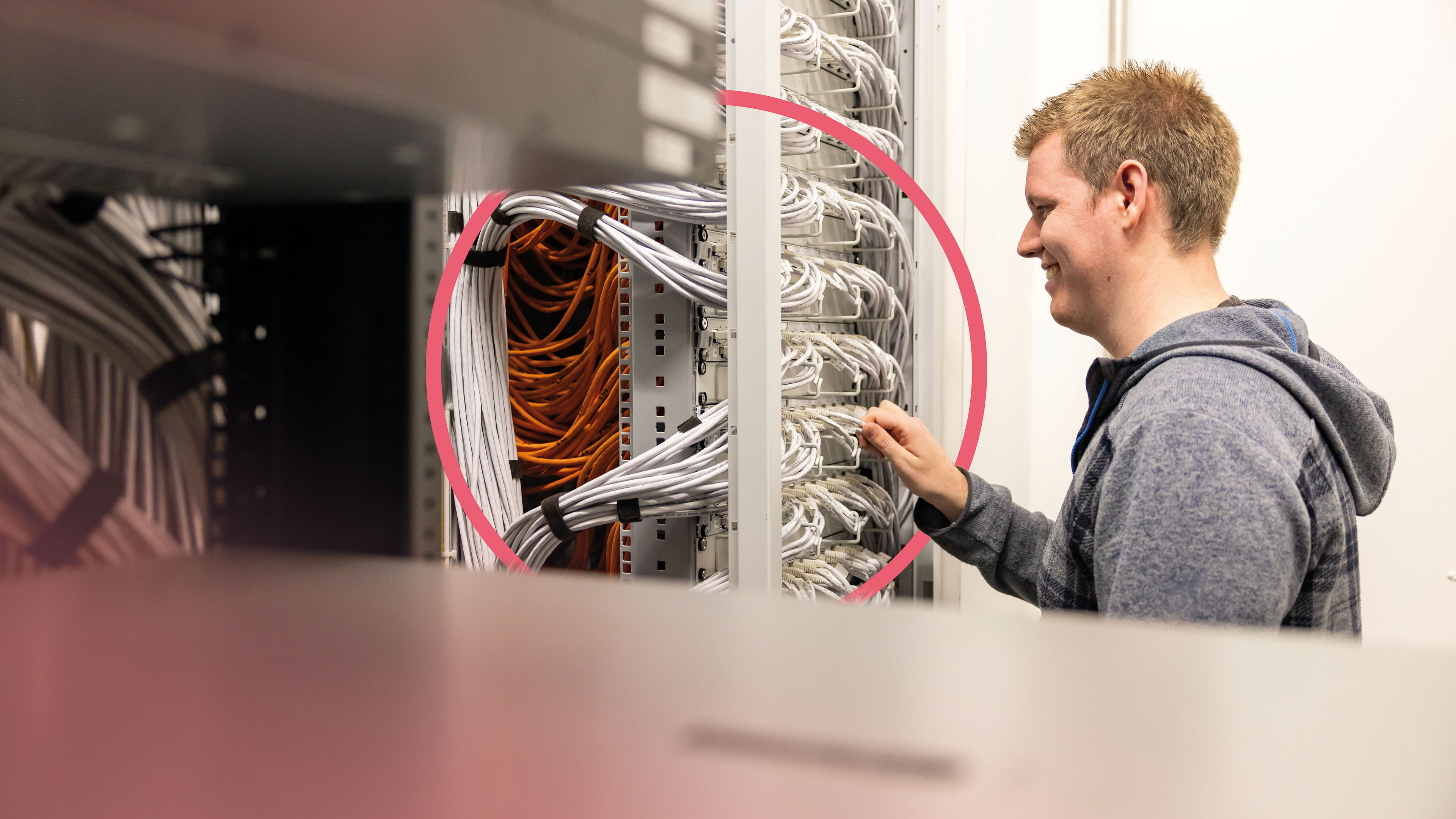 Mitarbeiter führt eine Anpassung am Ethernet-Patchpanel durch. Über dem Panel ist ein pinker Kreis als Gestaltungselement zu sehen.
