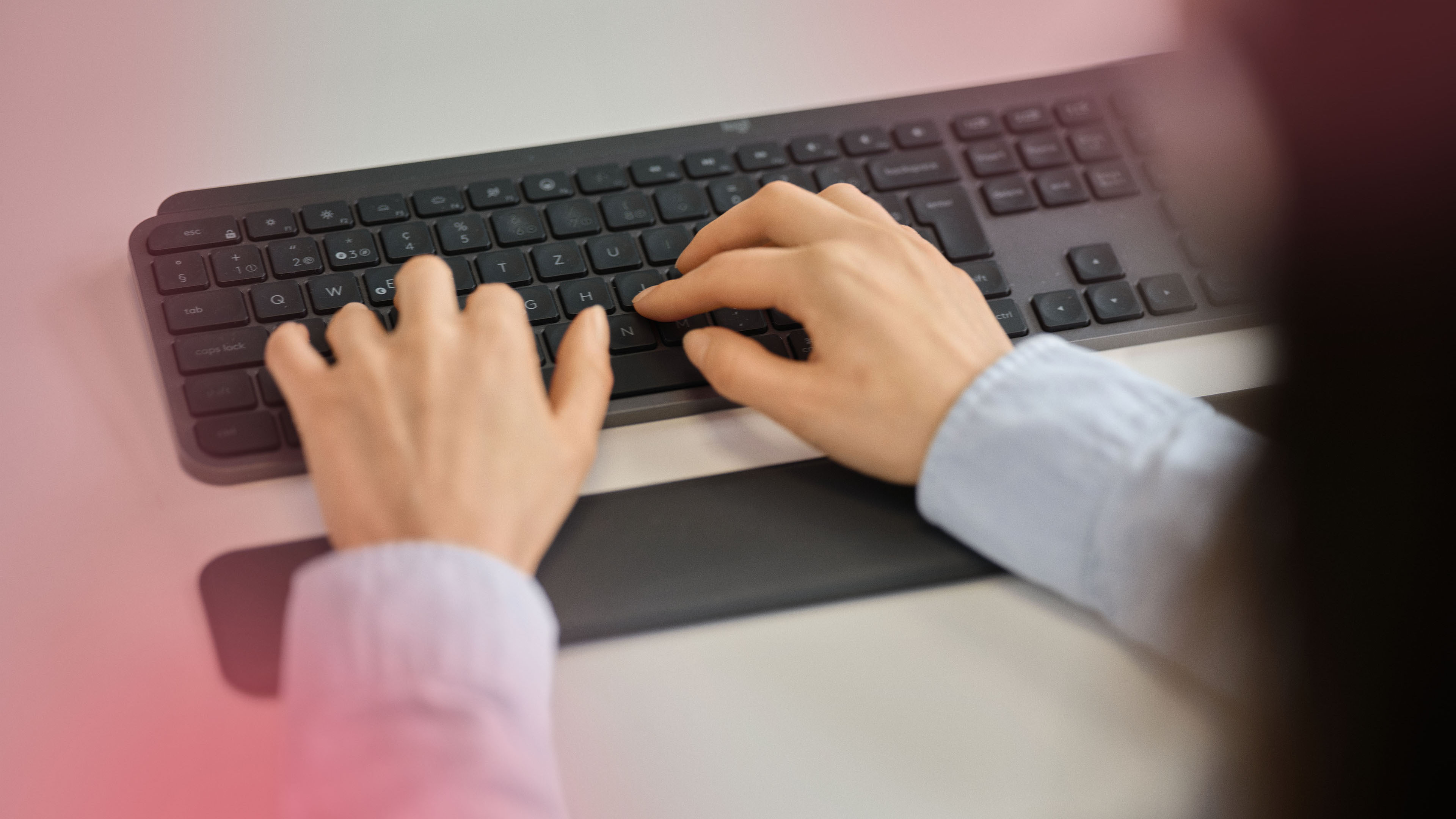 Nahaufnahme von zwei Händen beim Schreiben auf einer schwarzen Tastatur. Am Bildrand sind pinke Farbverläufe sichtbar.
