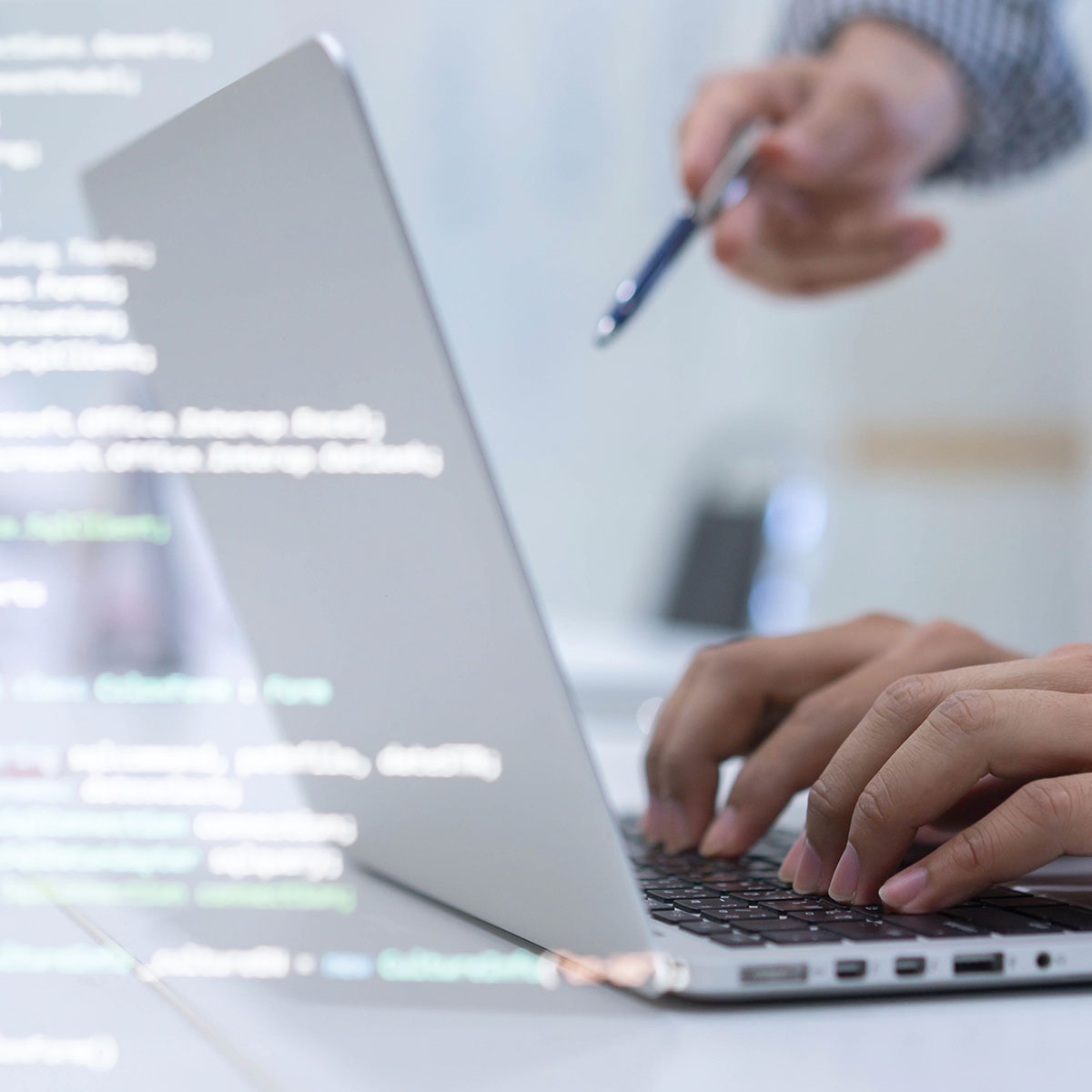 Hände tippen auf einer Tastatur, während Code als transparentes Overlay über dem Bild liegt. Im Hintergrund zeigt eine Person mit einem Stift auf den Bildschirm.