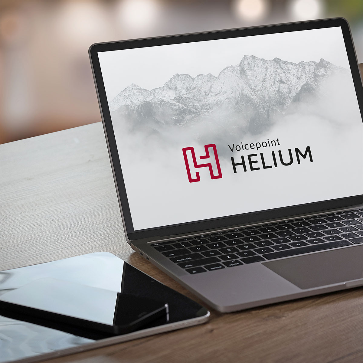 Ein Laptop zeigt das Logo von Voicepoint Helium, im Hintergrund sind Berge zu sehen. Daneben liegt ein Tablet auf dem Tisch.