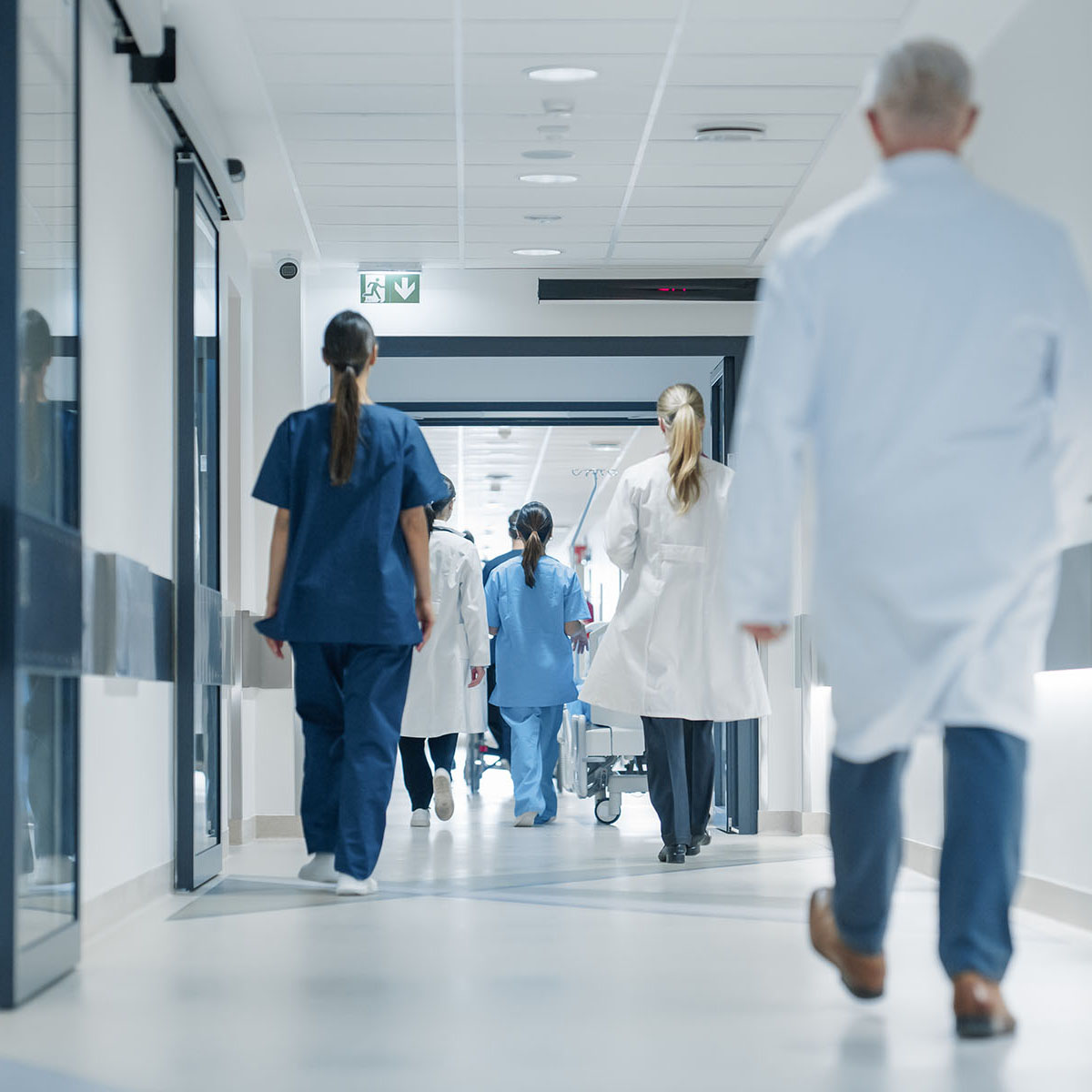 Mehrere Ärztinnen, Ärzte und Pflegefachpersonen gehen einen hellen Krankenhausflur entlang, einige tragen Laborkittel, andere blaue Arbeitskleidung.