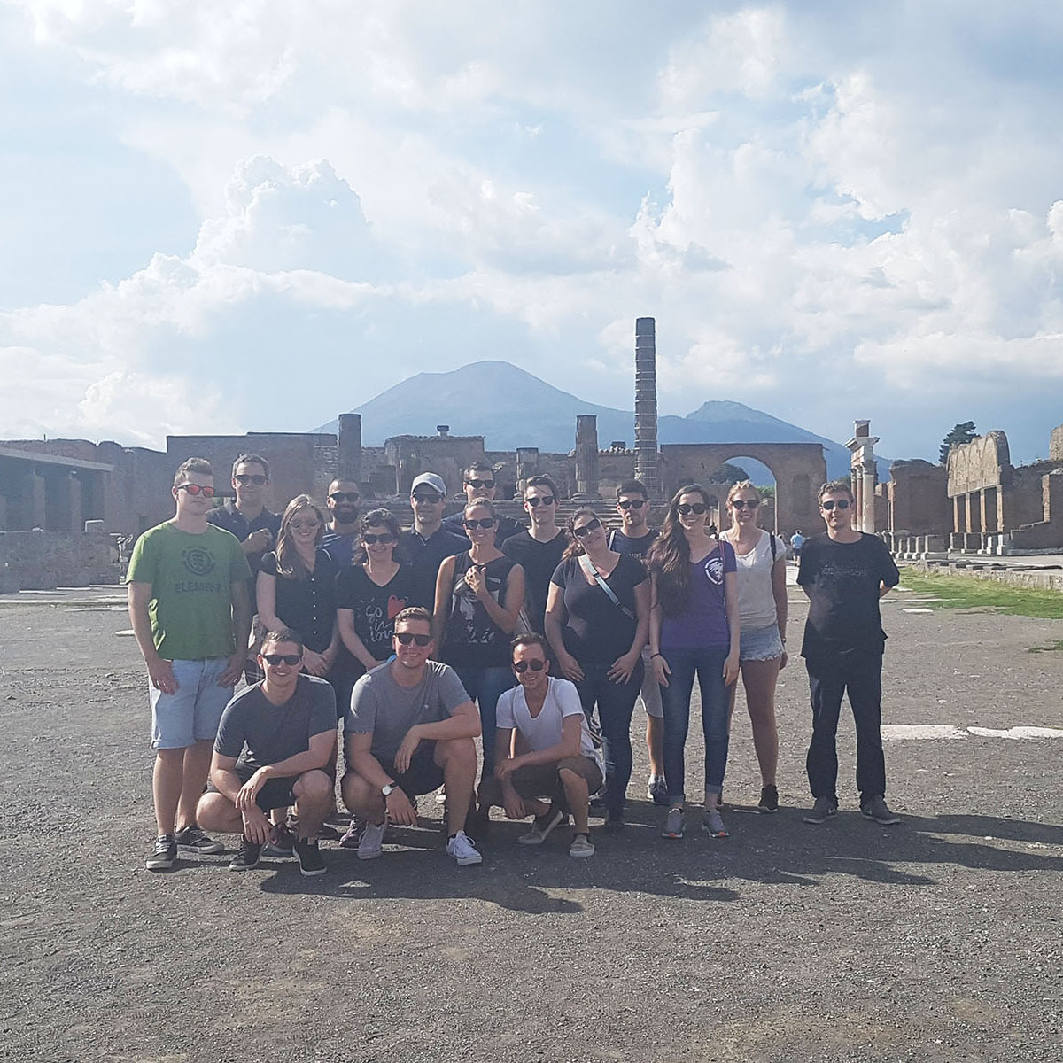 Gruppenfoto des Voicepoint-Teams vor einer historischen Ruinenstätte bei sonnigem Wetter, im Hintergrund sind Berge und blauer Himmel zu sehen.
