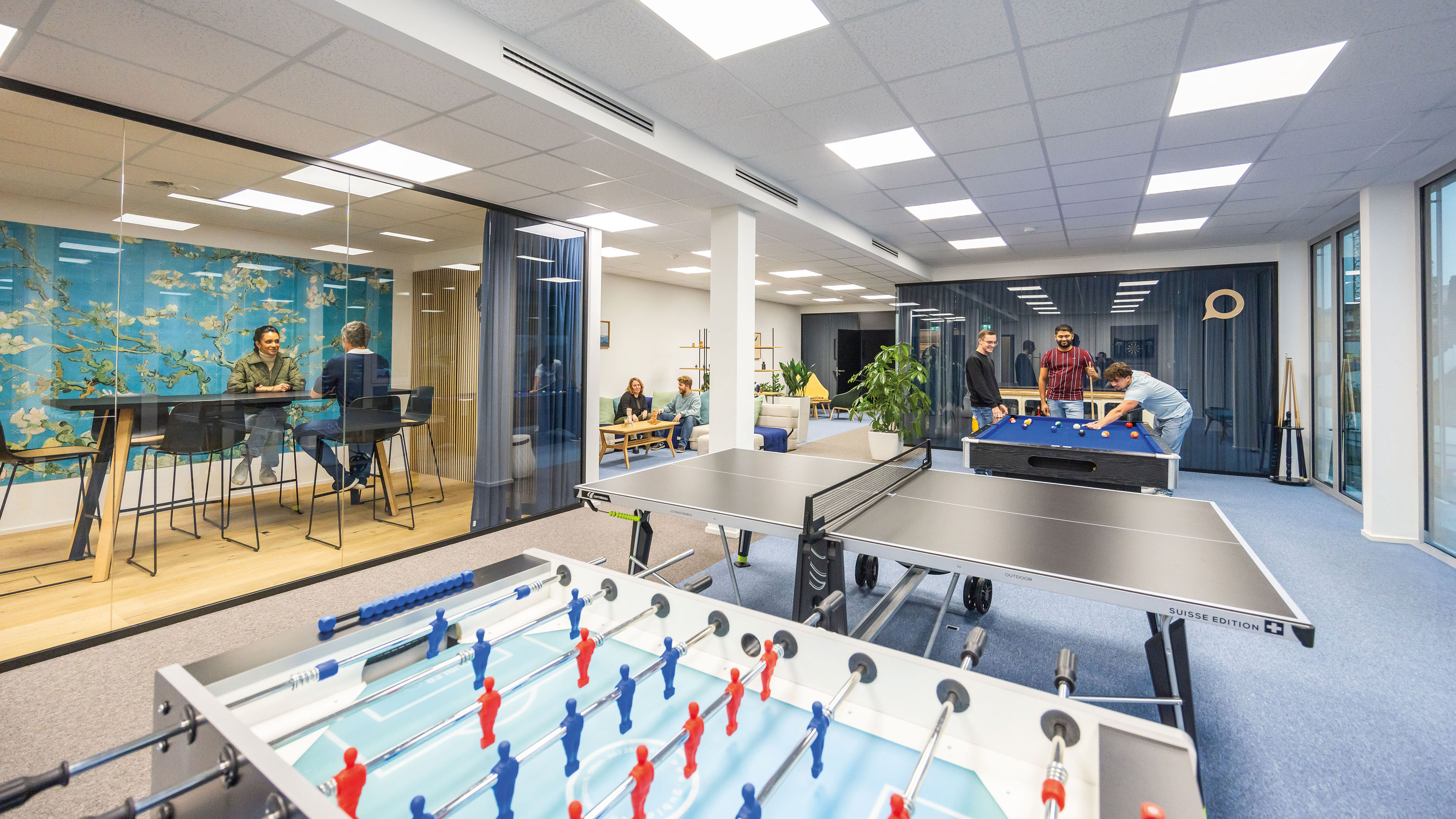Mitarbeitende spielen Billard und sitzen zusammen im Aufenthaltsbereich, vorne sind ein Pingpongtisch und Töggelikasten zu sehen.