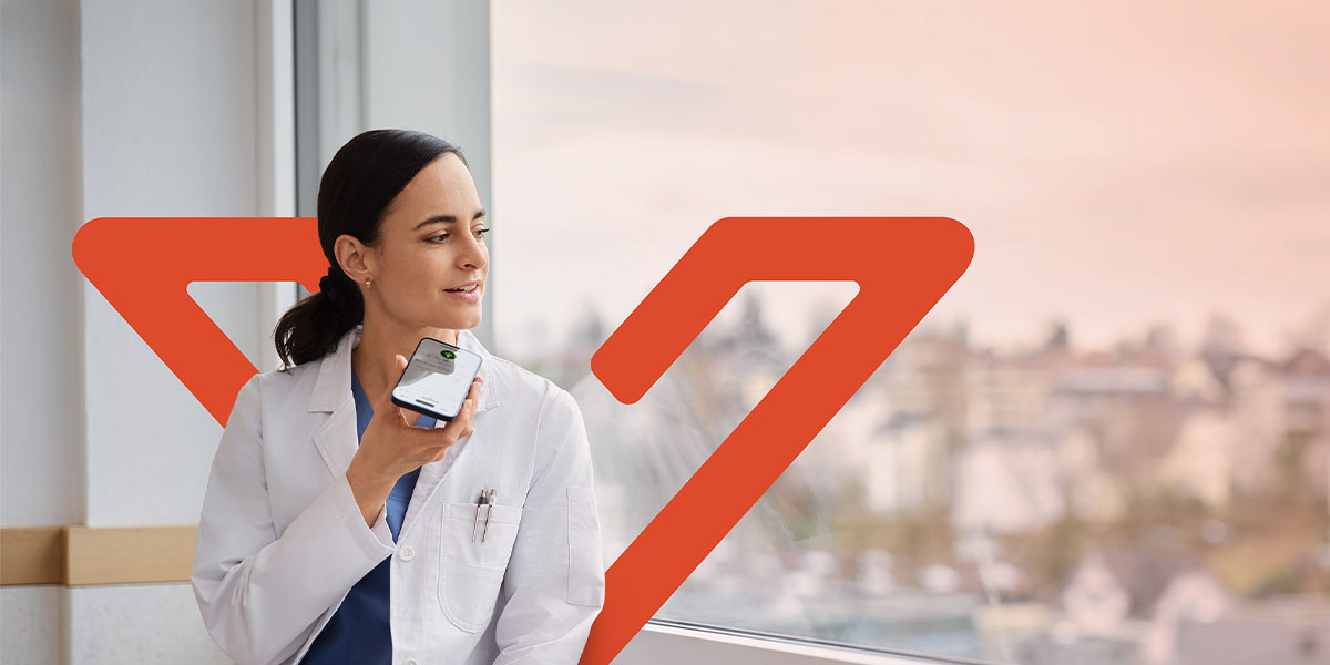 Ärztin sitzt vor einem Fenster und diktiert mit einem Smartphone, im Hintergrund ist das orange Voicepoint Xenon Logo sichtbar.