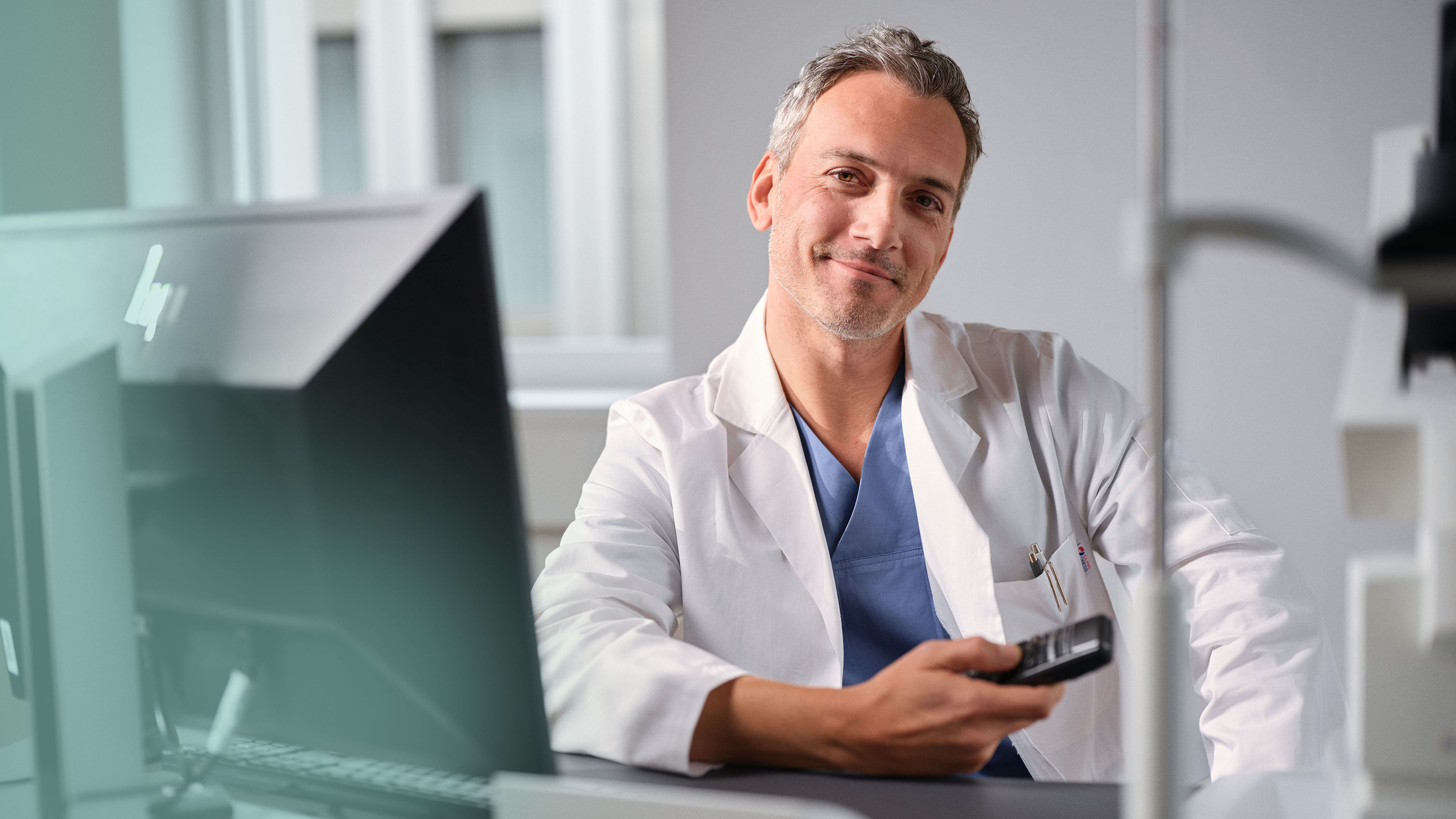 Arzt im weissen Kittel sitzt am Arbeitsplatz vor einem Windows Computer und diktiert mit Dragon Medical One.