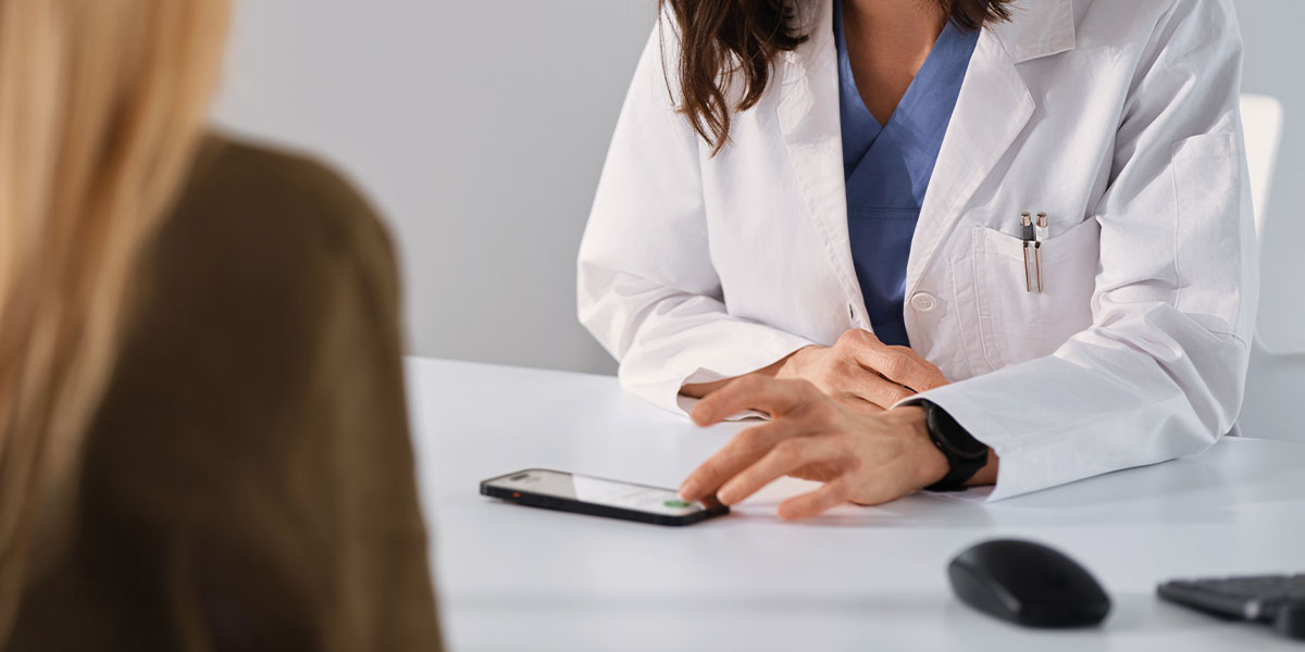 Ärztin sitzt in einem Behandlungszimmer der Patientin gegenüber und verwendet eine KI Lösung zur Aufnahme der Sprechstunde.