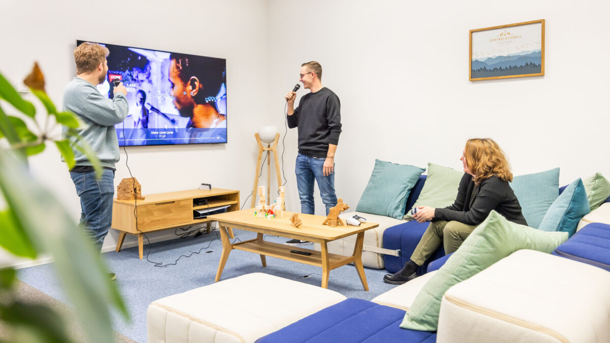 Zwei Mitarbeitende stehen im Aufenthaltsraum des Voicepoint-Büros in Wetzikon und singen Karaoke vor einem Fernseher.