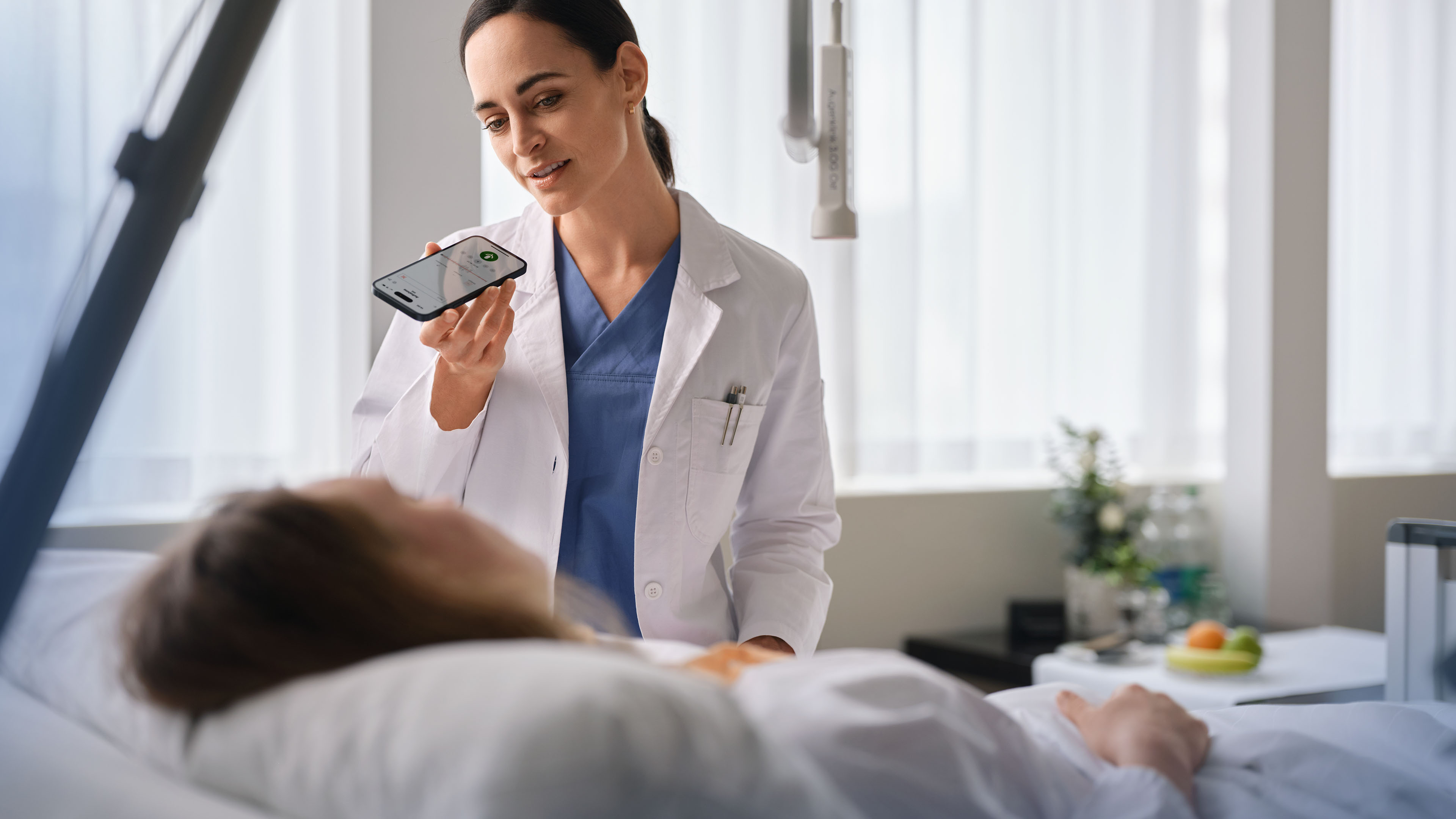 Ärztin steht am Patientenbett, spricht in ihr Smartphone und dokumentiert die Visite mit Voicepoint Xenon.