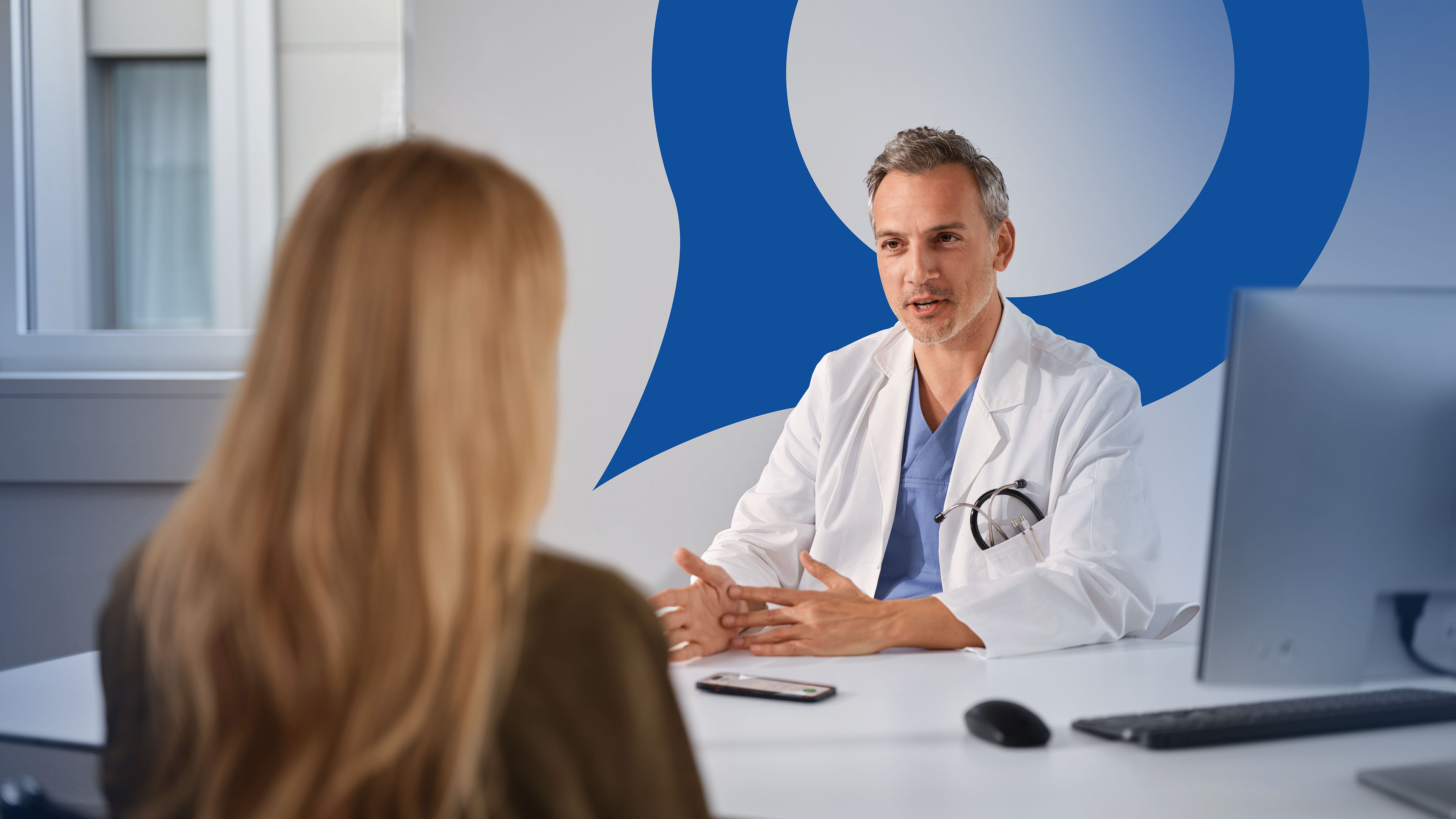 Arzt im Behandlungszimmer spricht mit Patientin, auf dem Tisch liegt sein Smartphone mit geöffneter Voicepoint Xenon App, die die Sprechstunde automatisch zusammenfasst.